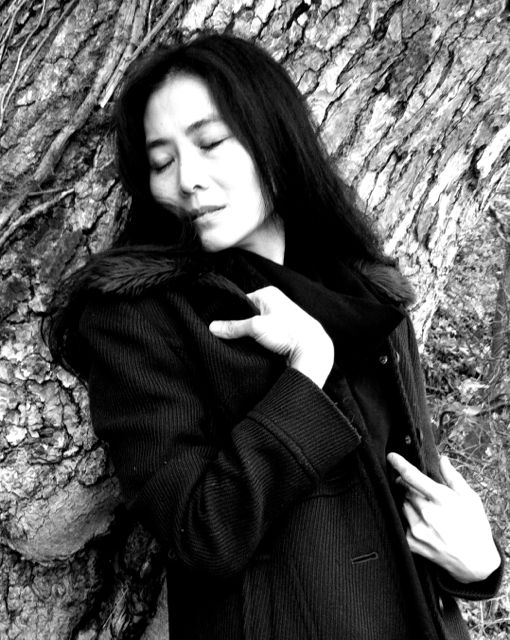 a black and white portrait of Mana Hashimoto leaning against the bark of a big tree, her eyes are closed and her hands cling to her jacket, it is as if the tree comforts her
